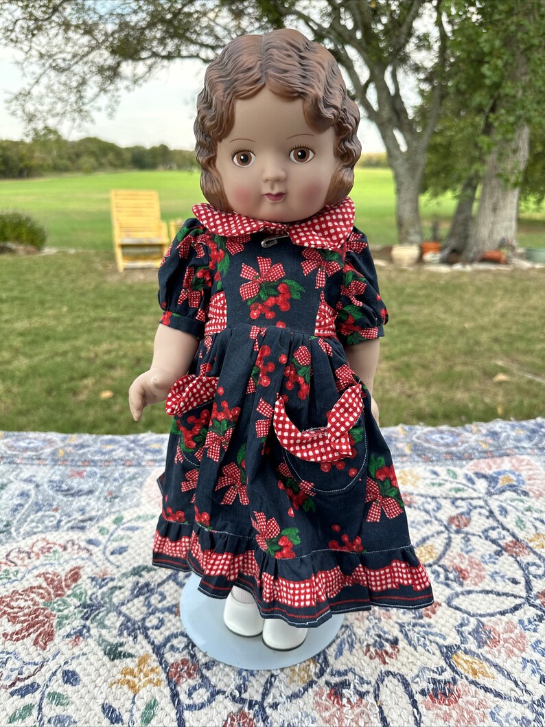 May include: A vintage doll with brown hair and eyes, wearing a navy blue dress adorned with red cherries and bows. The dress features a red and white checkered collar, pockets, and trim. The doll is wearing white shoes.