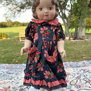 Könnte beinhalten: Eine Vintage-Puppe mit braunen Haaren und einem dunkelblauen Kleid, das mit roten Kirschen und Schleifen verziert ist. Das Kleid hat einen rot-weiß karierten Kragen, Taschen und Besatz. Die Puppe trägt weiße Schuhe und Socken.