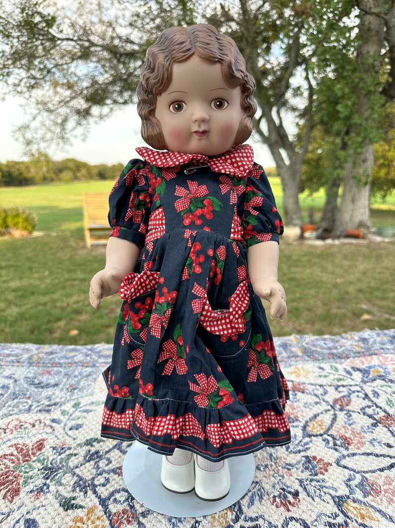 May include: A vintage doll with brown hair and a dark blue dress adorned with red cherries and bows. The dress features a red and white checkered collar, pockets, and trim. The doll is wearing white shoes and socks.
