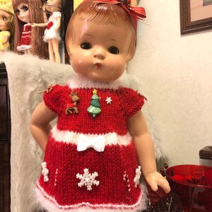 May include: A vintage doll in a handmade red and white Christmas dress, decorated with snowflake and candy cane details. The dress has a white bow and festive embellishments, including a reindeer and Christmas tree.