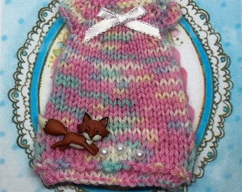 May include: Hand-knitted doll dress in pink, yellow, and blue yarn, adorned with a white bow and a brown fox button. The dress is embellished with three faux pearls. The text "Aeliza" and "Hand knitted for doll" are visible.
