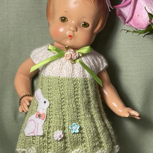 May include: A vintage doll wearing a handmade green and white knitted dress with a bunny applique and floral embellishments. The doll has a pink bow in her hair and a bracelet on her wrist. A pink rose is in the background.