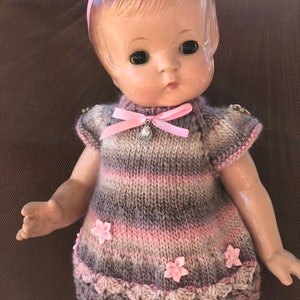 May include: A vintage doll with a pink ribbon headband and a knitted dress. The dress is striped in shades of gray and pink, adorned with pink flower accents and a scalloped hem. A pink bow and charm adorn the neckline.