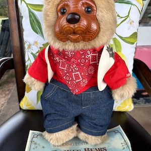 Może przedstawiać: Lalka misia Raikes Bears, edycja ślubna. Miś ma drewnianą twarz, brązowe futro i jest ubrany w czerwoną koszulę, dżinsowe szorty i czerwoną bandanę. Widoczny jest certyfikat autentyczności.