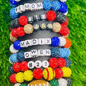 May include: A stack of colorful beaded bracelets with different sports-themed charms, including baseballs, footballs, and basketballs. The bracelets also feature letter beads spelling out names like "Jax", "Owen", "Maddix", and "Baylee".