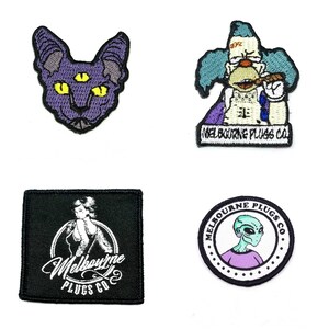 May include: Four embroidered patches with unique designs. Includes a purple cat with three eyes, a cartoon clown smoking a cigar, a woman in a circle, and an alien. All patches feature the text "Melbourne Plugs Co."