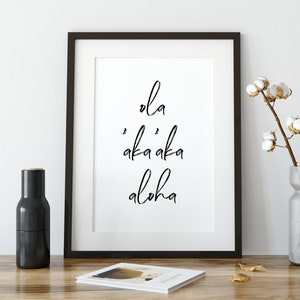 May include: Black and white framed print with the text "ola 'aka'aka aloha" on a white background. The print is sitting on a wooden table with a vase of cotton flowers and a black pepper grinder.