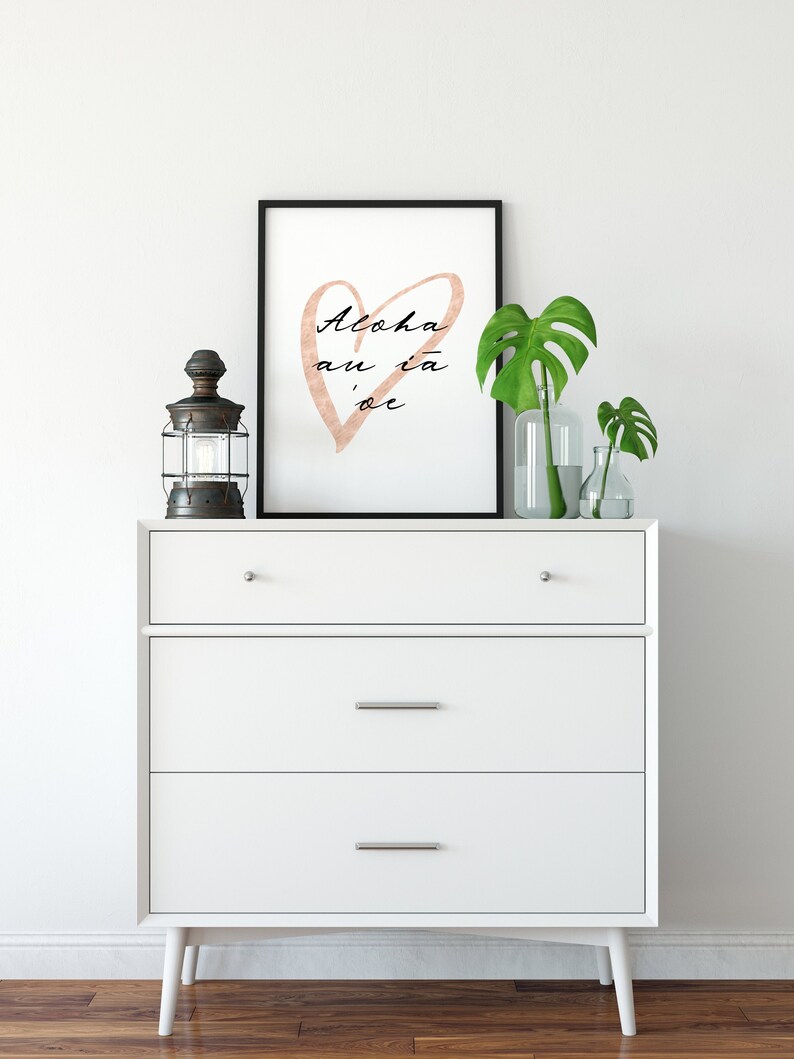 May include: White dresser with three drawers and black metal handles. A framed print with a pink heart and the text "Aloha au ia oe" is on top of the dresser. A black lantern and two glass vases with green plants are also on top of the dresser.