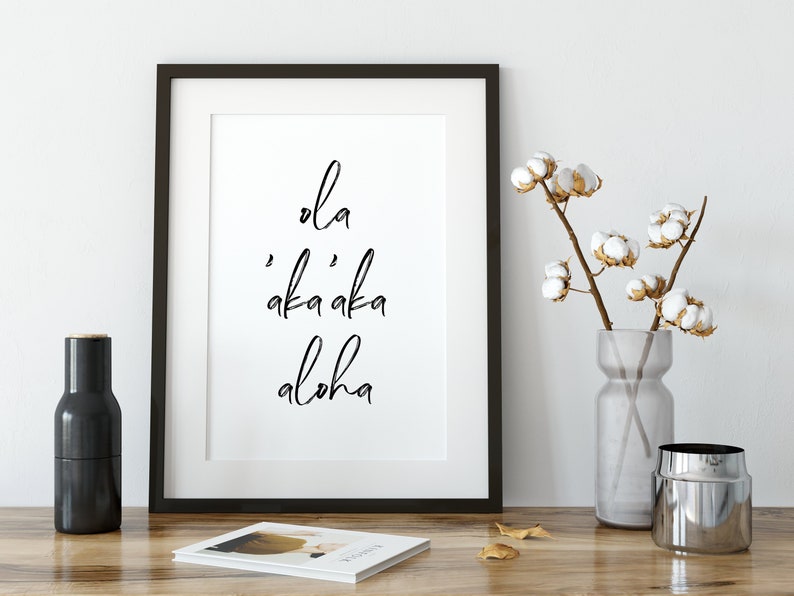 May include: Black and white framed print with the text "ola 'aka'aka aloha" on a white background. The print is sitting on a wooden table with a vase of cotton flowers and a black pepper grinder.