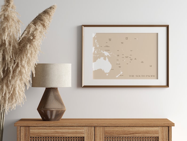May include: A framed map of the South Pacific with a beige background and white islands and text. The map is hanging on a white wall above a wooden dresser with a lamp on top.