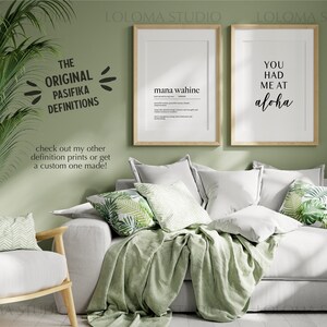 May include: Two framed prints with black text on a white background. The print on the left says "mana wahine" and the print on the right says "You had me at aloha". The prints are hanging on a sage green wall above a white couch with green throw pillows and a green throw blanket.