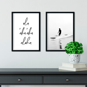 May include: Two black framed prints on a white wall. The print on the left has the text "'ola 'aka'aka aloha" in black script. The print on the right shows a black and white photo of a surfer riding a wave.