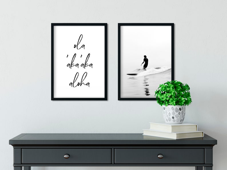 May include: Two black framed prints on a white wall. The print on the left has the text "'ola 'aka'aka aloha" in black script. The print on the right shows a black and white photo of a surfer riding a wave.