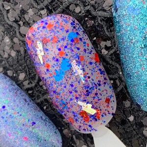 May include: Close-up of a fingernail with a clear base coat and red, blue, and silver glitter. The glitter is scattered randomly across the nail.
