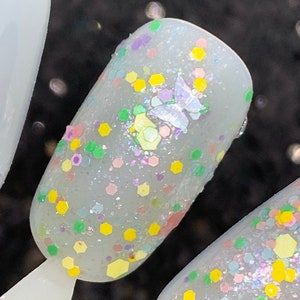 May include: Close-up of a light blue nail polish with a variety of colorful glitter, including yellow, green, pink, and purple. A small iridescent butterfly charm is also visible on the nail.
