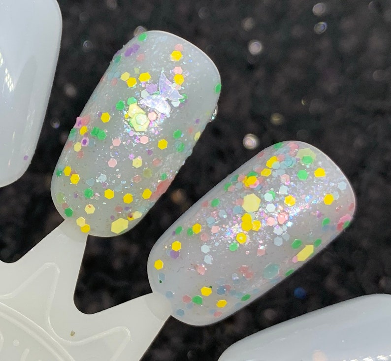 May include: Two light gray nail polish swatches with a variety of small, colorful glitter. The glitter is in shades of yellow, green, pink, and blue.