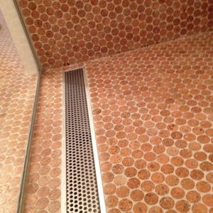 Op de afbeelding: Een close-up van een badkamer vloer met een afvoer gemaakt van een metalen rooster. De vloer is betegeld met kleine, ronde kurk tegels in een roodbruine kleur.