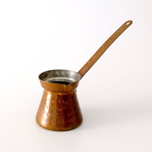 Könnte beinhalten: Eine kleine, kupferne Kaffeekanne mit gehämmerter Oberfläche und einem langen Griff. Die Kanne ist leer und steht auf einer weißen Oberfläche.