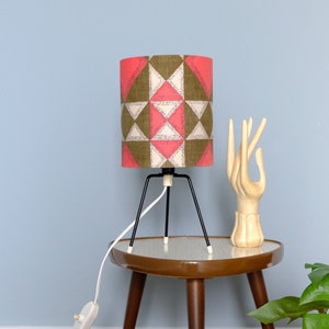 May include: A table lamp with a geometric patterned lampshade in shades of pink, brown, and white. The lamp has a black metal base with three legs and a white cord. The lamp is sitting on a brown table with a white top.
