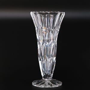 May include: A clear, crystal vase with a flared top and a detailed, textured design. The vase has a round base and a long, slender body. The vase is approximately 25 cm tall and is set against a black background.