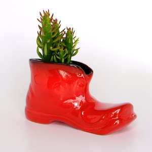 Könnte beinhalten: Ein roter Keramik-Stiefel-Pflanztopf mit einer grünen Sukkulentenpflanze darin.