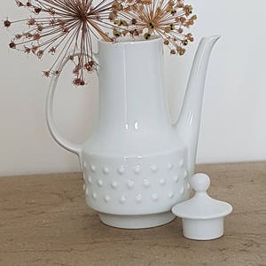 May include: White ceramic coffee pot with a textured design and a matching lid. The pot has a rounded body and a spout. The lid has a knob on top. The pot is sitting on a wooden surface.