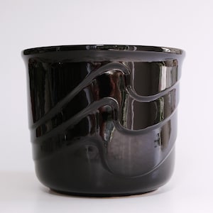 Könnte beinhalten: Ein schwarzer Keramiktopf mit einem wellenförmigen Design. Der Topf hat eine glänzende Oberfläche.