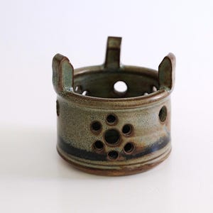 May include: A handmade ceramic candle holder with a cylindrical shape. The holder is glazed in shades of green, brown, and blue, with a series of circular holes and a flower-like pattern. It has three handles and a sturdy base.