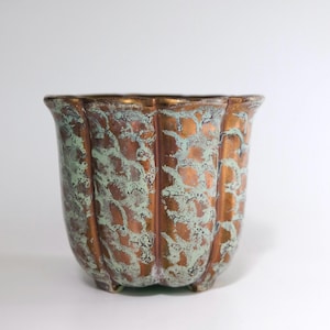 Op de afbeelding: Een decoratieve, kleine, koperkleurige plantenbak met een getextureerd oppervlak en een geschulpte rand. De plantenbak heeft een gevlekte groen-bruine patina en rust op drie kleine voetjes. De algemene vorm is een afgeronde vierkant.