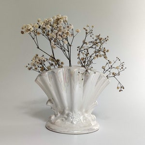 May include: White ceramic vase shaped like a seashell with a ruffled edge. The vase is filled with dried white flowers.