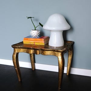 Puede incluir: Una mesa auxiliar pintada de oro con una lámpara en forma de hongo y una pequeña planta en maceta. La lámpara tiene una pantalla de vidrio blanco y una base blanca. La mesa tiene un diseño tallado y tres patas.