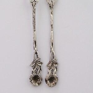 May include: A pair of silver teaspoons with ornate floral designs. The handles are intricately detailed with leaves and flowers. The back of each spoon is stamped with "925" indicating sterling silver.