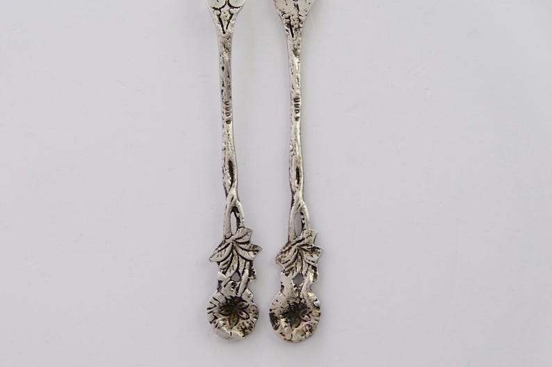 May include: A pair of silver teaspoons with ornate floral designs. The handles are intricately detailed with leaves and flowers. The back of each spoon is stamped with "925" indicating sterling silver.