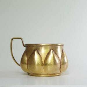 Könnte beinhalten: Eine goldfarbene Metalltasse mit Henkel. Die Tasse hat ein einzigartiges Design mit abgerundeten, blütenblattartigen Formen um den Körper. Der Henkel ist rechteckig und an der Seite der Tasse befestigt.
