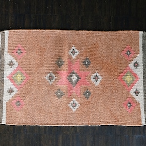 May include: A pink and white patterned rug with fringe on all sides. The rug has a geometric design with a central diamond shape.