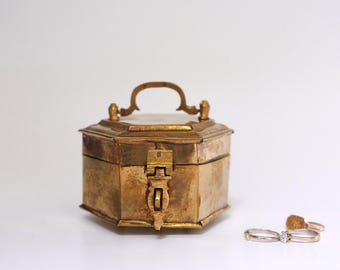 Puede incluir: Una caja de latón con tapa abatible y asa decorativa. La caja tiene forma hexagonal y un interior blanco. Dos anillos de oro están en primer plano.