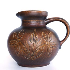 Puede incluir: Una jarra de cerámica marrón con asa y boquilla. La jarra presenta un diseño floral en relieve con dos flores y una planta central. Es un artículo decorativo.