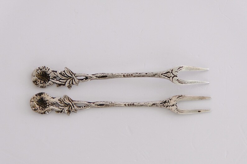 May include: A pair of silver-plated serving forks with ornate floral designs. The forks have two tines and are marked with "800" on the back.