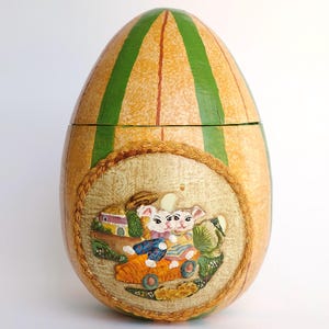 Puede incluir: Una caja decorativa con forma de huevo de Pascua con un exterior a rayas verde y amarillo. La caja está abierta para revelar una escena de dos conejos en un coche con una casa en el fondo.