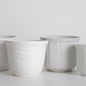 Può includere: Quattro vasi di fiori in ceramica bianca di diverse dimensioni e design. I vasi sono tutti di colore bianco semplice e hanno una finitura liscia.