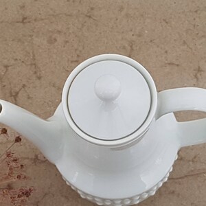 May include: White ceramic teapot with a lid and a handle. The teapot has a textured surface and a round, knob-like lid.