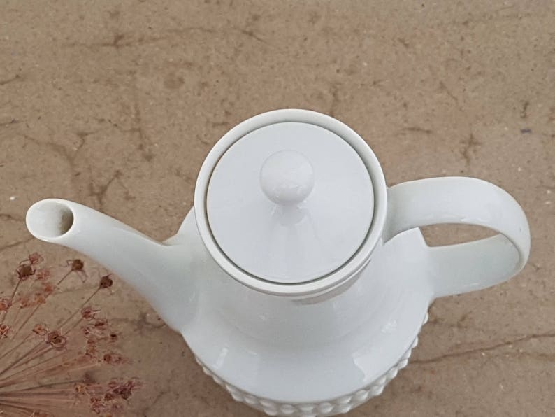 May include: White ceramic teapot with a lid and a handle. The teapot has a textured surface and a round, knob-like lid.