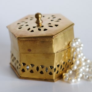 Op de afbeelding: Een gouden zeshoekige doos met ingewikkelde uitsnijdingen en een parelketting eroverheen gelegd.