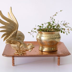 May include: A gold rooster figurine with outstretched wings sits on a hammered copper tray with three legs. A small potted plant with green leaves sits on the tray.