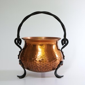 May include: A copper cauldron with a hammered texture and a black, twisted handle. The cauldron has three curved black legs and decorative black accents. The copper pot is a decorative item.