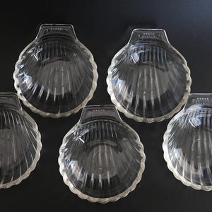 May include: Five clear glass seashell-shaped dishes. Each dish has a scalloped edge and a ribbed interior, resembling a real seashell. The dishes are arranged on a dark surface, showcasing their transparent quality.