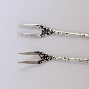 May include: Two silver-toned forks with floral designs. The forks have a twisted stem and a decorative floral design at the top.