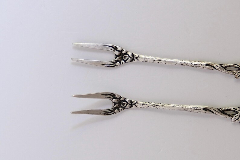 May include: Two silver-toned forks with floral designs. The forks have a twisted stem and a decorative floral design at the top.