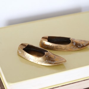 May include: Two gold-colored metal slipper-shaped ashtrays with intricate designs on a stack of books.