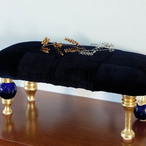 Op de afbeelding: Een kleine, donkerblauwe fluwelen voetenbank met goud geverfde poten en blauwe glazen eindstukken. De voetenbank is versierd met een gestoffeerd ontwerp.